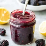A glass jar filled with homemade blackberry jam, surrounded by fresh blackberries and lemon halves—perfect for easy jam recipes and fruit preserves.