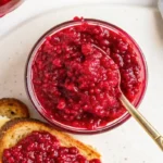 Homemade raspberry jam in a mason jar with spoon on a marble countertop