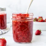 Homemade strawberry freezer jam in a glass jar with a spoon, surrounded by fresh strawberries on a white background.