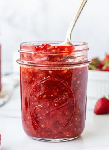 Homemade strawberry freezer jam in a glass jar with a spoon, surrounded by fresh strawberries on a white background.