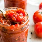 Homemade tomato relish in a glass jar with a spoonful, surrounded by fresh tomatoes