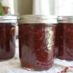Homemade strawberry jam in glass canning jars with fresh strawberries and sugar on rustic wooden background