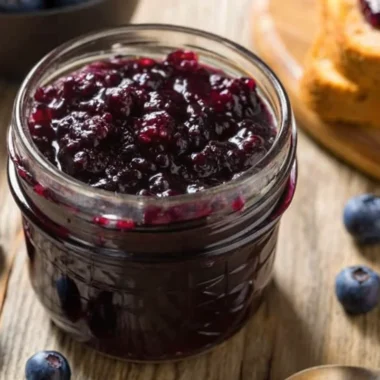 Fresh huckleberry jam in glass jars with whole huckleberries scattered around, showing the deep purple color and smooth texture of this homemade huckleberry jam recipe
