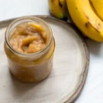 Golden homemade banana jam in mason jar with wooden spoon showing thick, glossy texture against rustic kitchen backdrop