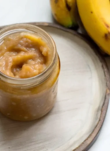 Golden homemade banana jam in mason jar with wooden spoon showing thick, glossy texture against rustic kitchen backdrop