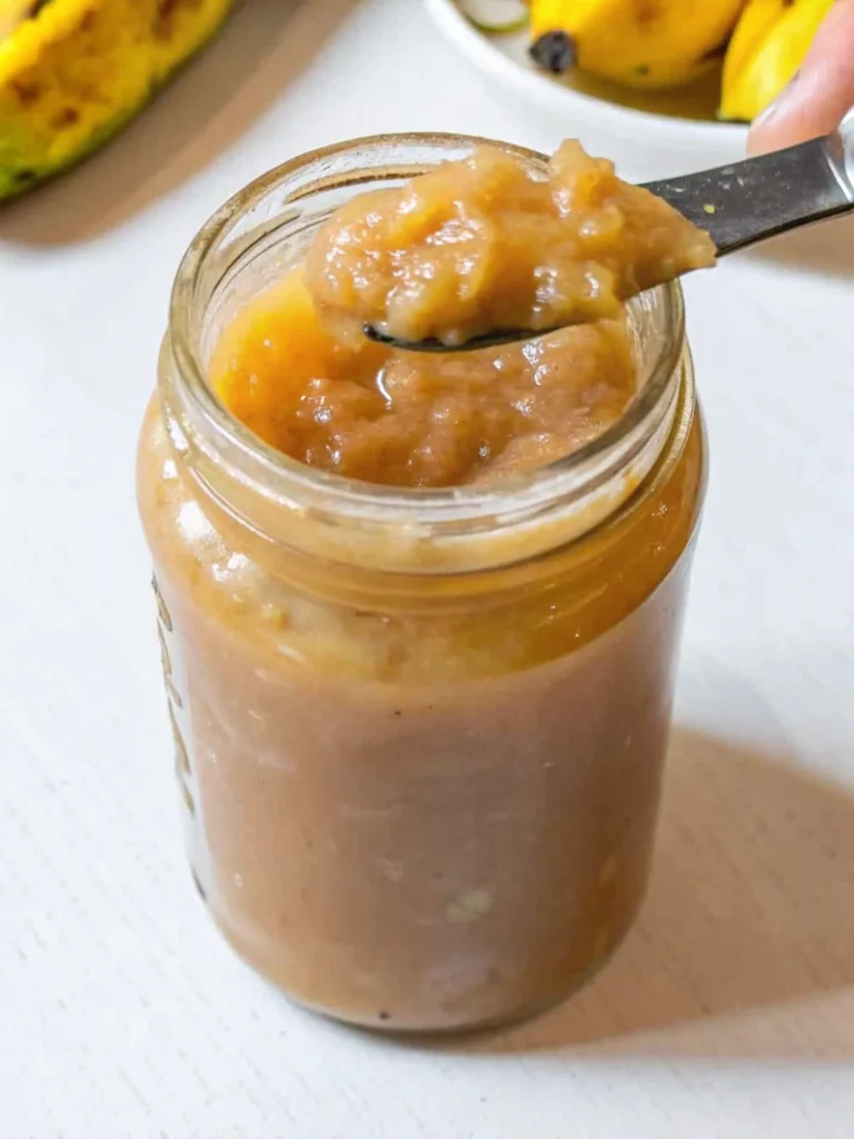 Golden homemade banana jam in mason jar with wooden spoon showing thick, glossy texture against rustic kitchen backdrop