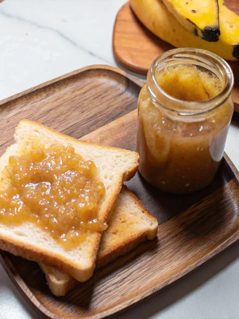 Golden homemade banana jam in mason jar with wooden spoon showing thick, glossy texture against rustic kitchen backdrop