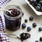 Homemade blueberry jam without pectin in glass jar with fresh blueberries scattered around wooden cutting board
