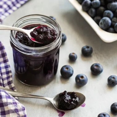 Homemade blueberry jam without pectin in glass jar with fresh blueberries scattered around wooden cutting board