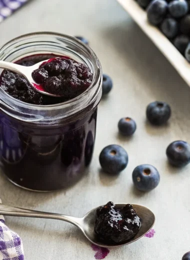 Homemade blueberry jam without pectin in glass jar with fresh blueberries scattered around wooden cutting board