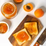 Fresh homemade apricot jam in sterilized glass jars with golden orange color, showing thick consistency and small fruit pieces, placed on rustic wooden surface with fresh apricot halves and vintage spoon for professional food photography