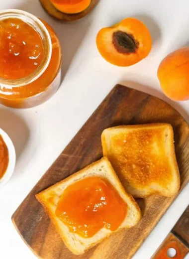 Fresh homemade apricot jam in sterilized glass jars with golden orange color, showing thick consistency and small fruit pieces, placed on rustic wooden surface with fresh apricot halves and vintage spoon for professional food photography