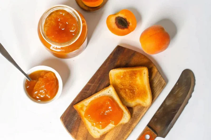 Fresh homemade apricot jam in sterilized glass jars with golden orange color, showing thick consistency and small fruit pieces, placed on rustic wooden surface with fresh apricot halves and vintage spoon for professional food photography