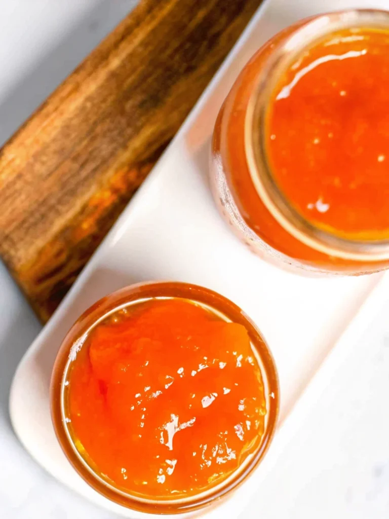Fresh homemade apricot jam in sterilized glass jars with golden orange color, showing thick consistency and small fruit pieces, placed on rustic wooden surface with fresh apricot halves and vintage spoon for professional food photography