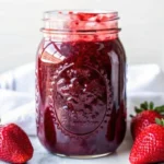 Homemade strawberry jam canning recipe showing fresh strawberries, sugar, and finished sealed jars of bright red strawberry preserves on wooden counter