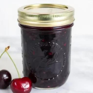 Homemade cherry jam recipe in glass jar with fresh cherries and wooden spoon on rustic kitchen counter