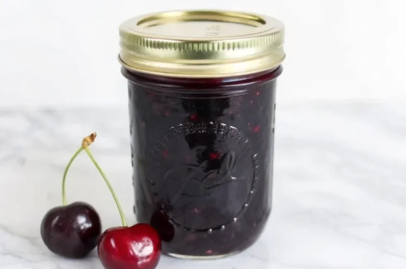 Homemade cherry jam recipe in glass jar with fresh cherries and wooden spoon on rustic kitchen counter