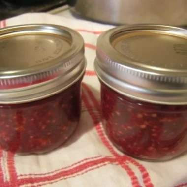 Homemade salmonberry jam recipe in mason jars with fresh salmonberries scattered around wooden cutting board, featuring golden-orange preserve with visible fruit pieces