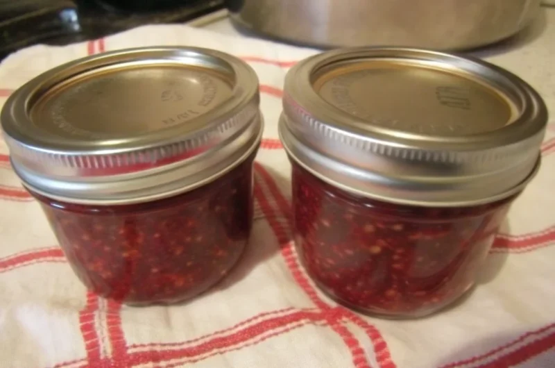 Homemade salmonberry jam recipe in mason jars with fresh salmonberries scattered around wooden cutting board, featuring golden-orange preserve with visible fruit pieces