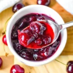 Homemade cherry jelly recipe no pectin in glass jar with fresh cherries scattered around wooden cutting board