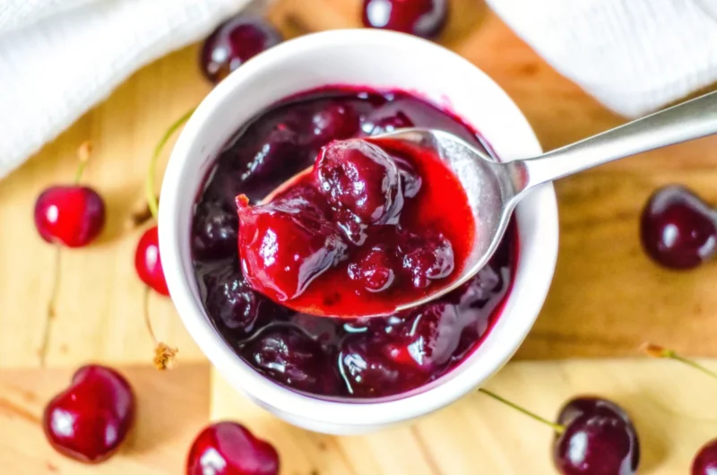 Homemade cherry jelly recipe no pectin in glass jar with fresh cherries scattered around wooden cutting board