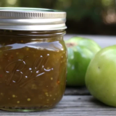 Homemade green tomato jam in glass jars with fresh green tomatoes and canning supplies on rustic kitchen counter