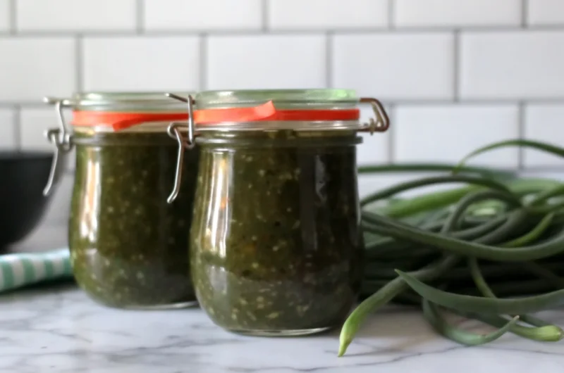 Homemade garlic scape jam in glass jars with fresh garlic scapes and herbs on rustic wooden table