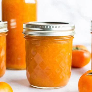 Homemade persimmon jam in glass jars with fresh fuyu persimmons, lemon slices, and wooden spoon on rustic kitchen counter