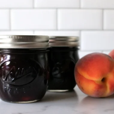 Homemade blueberry peach jam in glass mason jars with fresh peaches and blueberries scattered around, showing the beautiful ruby-red color of the finished preserve