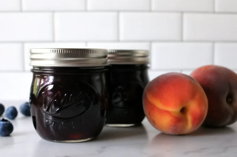 Homemade blueberry peach jam in glass mason jars with fresh peaches and blueberries scattered around, showing the beautiful ruby-red color of the finished preserve