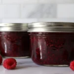 Homemade thimbleberry jam in glass jars with fresh thimbleberries scattered around wooden cutting board