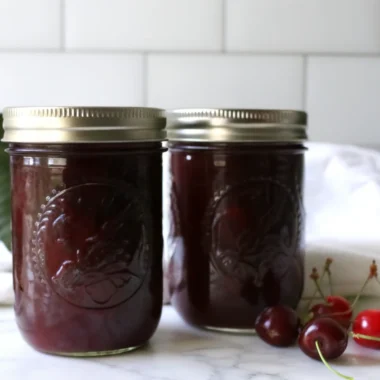 Homemade sour cherry jam in glass jars with fresh cherries scattered around on wooden cutting board